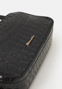 Borsa in ecopelle nera con texture a forma di coccodrillo, dettagli in oro con logo, forma rettangolare e finitura liscia. Chiusura con zip e manico piatto.