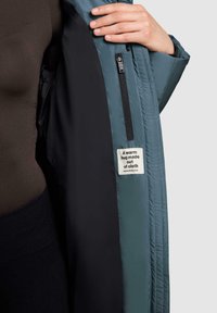 Hånd der åbner en teal farvet jakke, der viser sort indvendigt for, en lynlås lomme og et stofmærke med teksten "Et varmt kram lavet af stof."