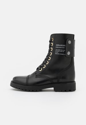 Schwarze Lederkampfstiefel mit goldenen Ösen, schwarzen Schnürsenkeln und einem Rückengurt mit gedrucktem Text. Dicke, strukturierte Gummisohle.