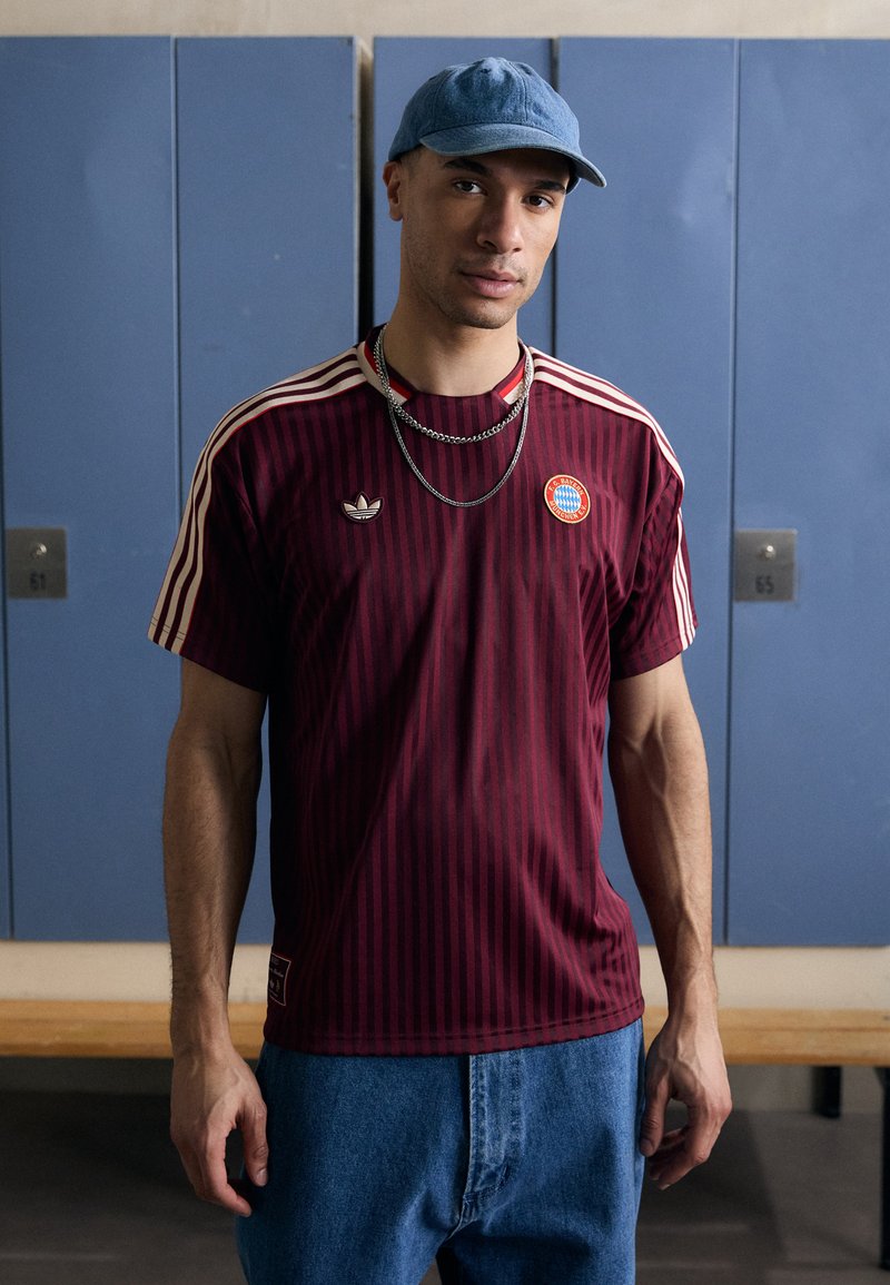 Maglietta Adidas a maniche corte a righe bordeaux con dettagli crema e patch del logo, abbinata a jeans in denim blu chiaro e un berretto blu chiaro.