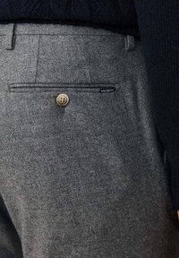 Sive teksturirane hlače s stražnjim džepom, koje imaju smeđi gumb i crnu oznaku s imenom branda "HACKETT."