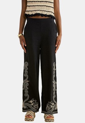 Pantalones negros de pierna ancha con bordado floral blanco a lo largo de los laterales y dobladillos, combinados con sandalias de plataforma beige tejidas y un top de crochet color crema.