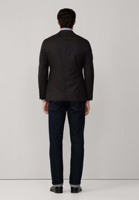 Blazer scuro da uomo abbinato a jeans scuri. Il blazer presenta una vestibilità su misura e una silhouette strutturata. Tessuto liscio, senza motivi visibili.