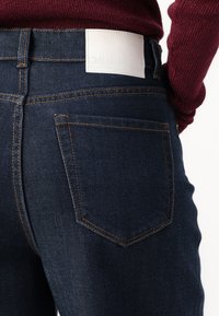 Jeans de mezclilla de un azul oscuro con costuras naranjas, que presentan un bolsillo trasero y una etiqueta de marca de cuero blanco en la cinturilla.