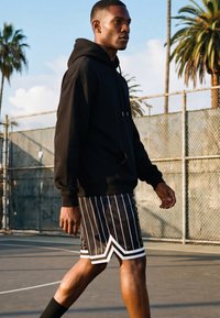 Jonge man in een zwarte hoodie en gestreepte korte broek loopt op een buitenbasketbalveld met palmbomen en een gaashek op de achtergrond.