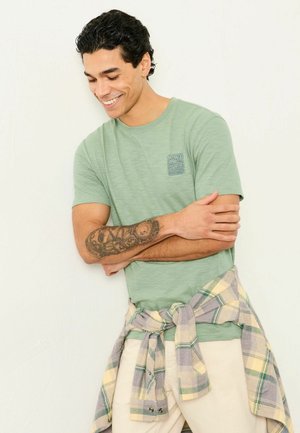 Light green t-shirt with a small blue logo, accompanied by a tied light plaid flannel shirt around the waist and beige pants.