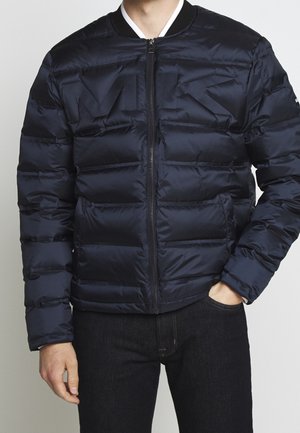 Homme portant une doudoune bleu foncé avec une fermeture éclair à l'avant, design matelassé, et un pantalon noir, debout avec une main dans la poche.