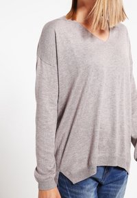 Femme portant un pull ample gris clair à col en V avec manches longues et un jean bleu, montrée du cou aux hanches sur un fond blanc.