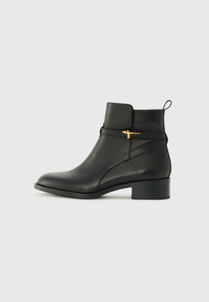 Bottine en cuir noir avec un low heel, dotée d'un accent doré et d'une languette à l'arrière. Texture lisse et design épuré.