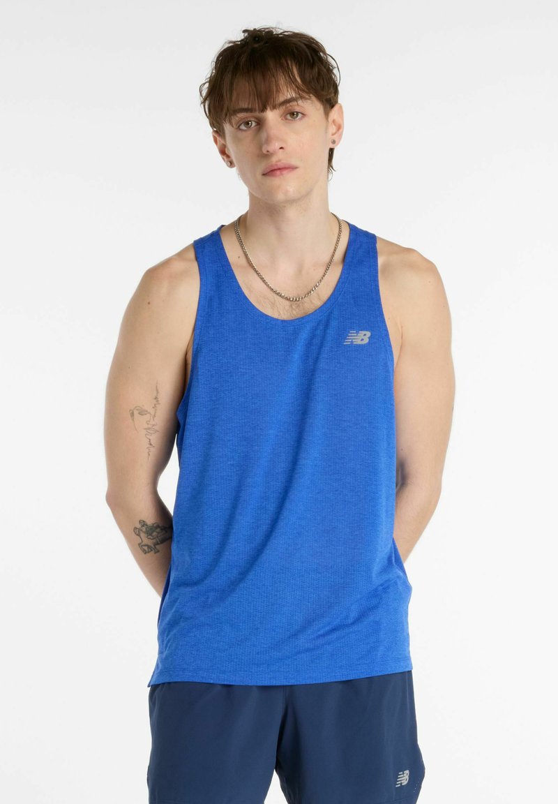 Junger Mann mit kurzen braunen Haaren, der ein blaues New Balance Tanktop und dunkle Shorts trägt, steht mit den Händen hinter dem Rücken vor weißem Hintergrund.