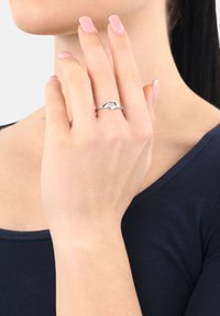 Mano con uñas rosadas pulidas tocando el cuello, usando un anillo de plata con un diseño en forma de corazón en el dedo medio, vestida con una blusa de color azul oscuro.