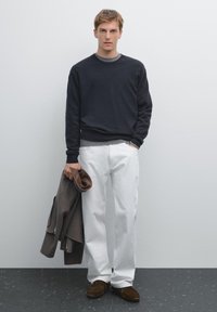 Marineblauer Pullover mit grauem Rundhalsausschnitt, weißen geraden Hosen und braunen Wildlederschuhen. Hält eine braune Jacke. Minimalistischer Hintergrund.