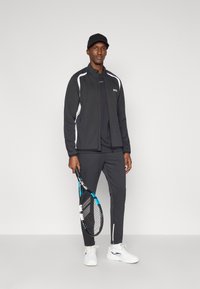 Μαύρο αθλητικό σακάκι με λευκές πλευρικές λεπτομέρειες, μαύρη μπλούζα, και ταιριαστά παντελόνια. Κρατάει μια ρακέτα τένις, φοράει λευκά αθλητικά παπούτσια και καπέλο.