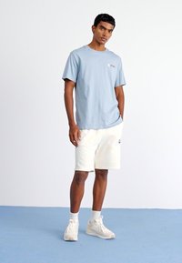 Blå bomulds T-shirt med rund hals, parret med hvide shorts. Outfittet har subtil branding og en afslappet pasform. Skoene er beige.