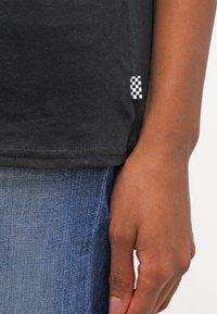 T-shirt en coton noir avec un ourlet droit, comportant une petite étiquette à damier noire et blanche sur le côté. Jean en denim bleu visible.