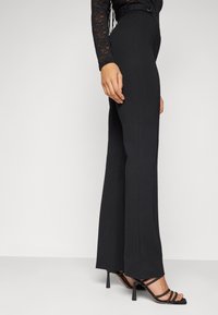 Black flared pants with a high waist, smooth fabric, and a button detail. Paired with black strappy heels, highlighting a slim silhouette.
