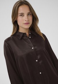 Dark brown satin button-up shirt with a pointed collar, long sleeves, and white buttons down the front, showcasing a smooth, glossy finish.