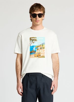 Mann trägt schwarze Sonnenbrille, weißes T-Shirt mit bunter Küstenillustration und Text sowie dunkle Hose vor schlicht hellem Hintergrund.