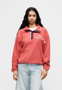 Γυναίκα που φοράει κοραλλί φλις πουλόβερ με κουμπιά τύπου snap και φερμουαριστή τσέπη στο στήθος, συνδυασμένο με ανοιχτό μπλε τζιν με σκισίματα, στέκεται μπροστά σε λευκό φόντο.