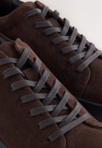 Baskets en daim marron avec lacets gris foncé, gros plan montrant la texture et les lacets entrelacés sur les deux chaussures.
