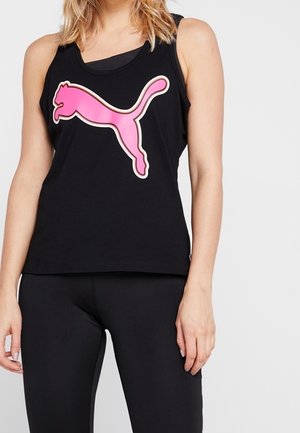 Kvinde iført sort ærmeløs top med stort pink Puma-logo og sorte leggings, stående mod en ensfarvet lys baggrund.