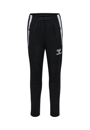 Smilende dreng med mørkt hår iført en gul og sort sportsjakke og sorte træningsbukser, der står med hænderne bag ryggen.