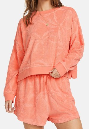 Femme portant un sweat-shirt en éponge corail et un short assorti avec un motif en relief de feuille de palmier et un collier pendentif en or.