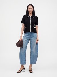 Femme debout, portant un haut noir en maille à manches courtes, un jean bleu ample, des sandales à talons noires et tenant un sac à main marron texturé.