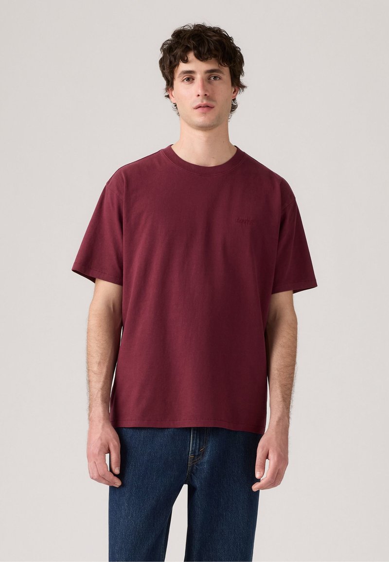 Camiseta de algodón burdeos de mangas cortas, con cuello redondo y un diseño simple. Presenta un branding minimalista en el pecho.