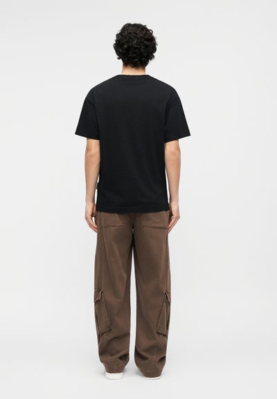 Person mit lockigen dunklen Haaren, trägt ein schlichtes schwarzes T-Shirt und lockere braune Cargo-Hosen, steht mit dem Rücken zum Betrachter vor einem weißen Hintergrund.