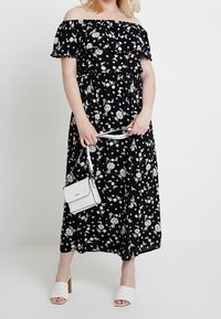 Czarna maksigłówkowa sukienka w kwiaty z dekoltem typu off-the-shoulder i falbanami, zestawiona z białymi sandałami na obcasie oraz małą białą torebką.