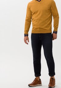 Pull col en V moutarde porté sur une chemise à carreaux bleu marine avec boutons, assorti à un jean foncé et des chaussures de sport marron avec semelles blanches et détails noirs.