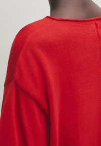 Haut en tissu rouge et doux avec un décolleté large et une coupe décontractée. Présente des coutures d'épaule discrètes et une texture lisse.