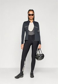 Žena u crnom biciklističkom odijelu drži crnu kacigu, nosi sunčane naočale i biciklističke cipele, stoji nasuprot jednostavne pozadine.