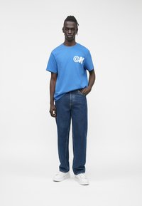 Μπλε βαμβακερό t-shirt με λευκό λογότυπο, σε συνδυασμό με φαρδιά μπλε τζιν τζιν και λευκά αθλητικά παπούτσια. Απλός και καθημερινός σχεδιασμός.