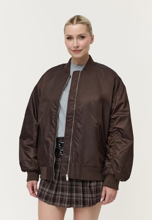 Kvinde iført en brun oversized bomberjakke, grå top og brun ternet nederdel, stående mod en ensfarvet lys baggrund.