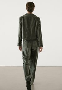 Dunkelgrüne, kurze Jacke mit glatter Textur und zwei Knopfakzenten, kombiniert mit passenden Hosen, von hinten betrachtet.