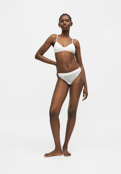 Jeune femme debout pieds nus, portant un soutien-gorge blanc et un sous-vêtement en résille sur un fond clair uni, une main sur la hanche.