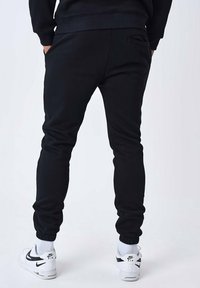 Zwarte sweatpants van zacht materiaal met geribde manchetten, voorzien van een achterzak en gecombineerd met witte sneakers en zichtbare geribde enkelsokken.