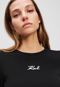 Svart ribbestrikket genser med rund hals og hvit brodert "Karl"-logo på brystet, med glatt stoffstruktur og tettsittende design.