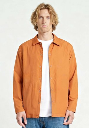 Jonge man met halflang blond haar, een oranje jack met knopen over een wit shirt en blauwe spijkerbroek, tegen een effen achtergrond.
