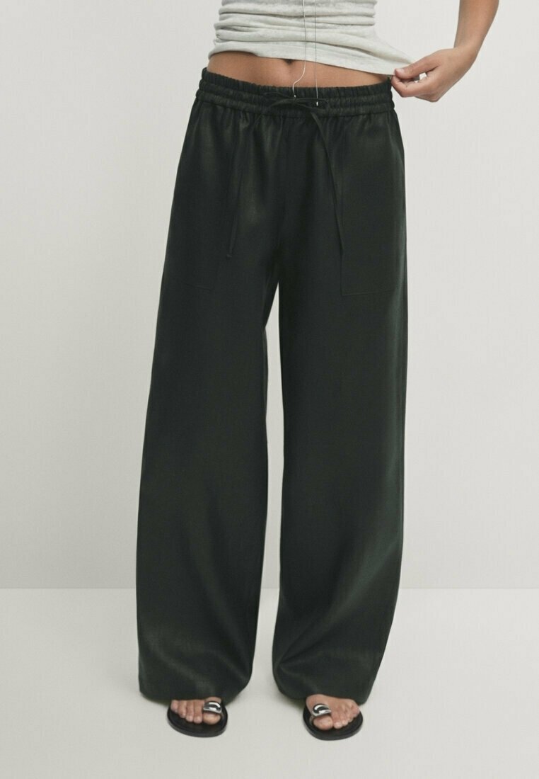 Femme portant un pantalon large noir et ample avec une taille à cordon et des sandales noires, debout devant un fond clair uni.