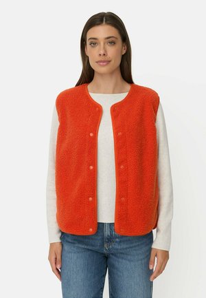 Femme portant une veste polaire orange boutonnée par-dessus une chemise blanche à manches longues et un jean bleu, debout devant un fond uni.