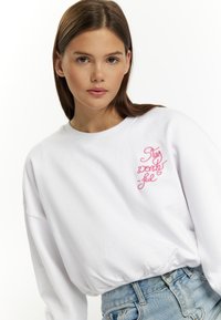 Witte sweatshirt met lange mouwen en ronde hals, met roze geborduurde tekst "Blijf geweldig" op de borst. Draagt met blauwe spijkerbroek shorts.