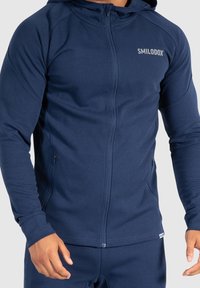 Sudadera con cremallera en color navy, fabricada en un tejido suave con una textura lisa. Cuenta con capucha, bolsillos laterales y el logo "SMILODOX" en el pecho.