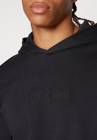 Čierna mikina s kapucňou s textúrovanou tkaninou. Obsahuje diskrétne vyšívanie 'Calvin Klein' v strede. Detailný pohľad na oblasť krku a švy.