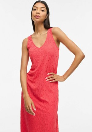 Femme portant une robe corail texturée sans manches avec un décolleté en V, debout, une main sur la hanche et l'autre relâchée sur le côté.