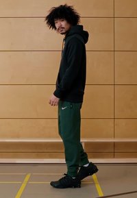 Juodas džemperis su gaubtu ir žaliais sportiniais kelnėmis kišenėse, su Nike logotipu. Juodi sportiniai batai su tekstūruota padais, stovintys patalpose prie medinių panelių.