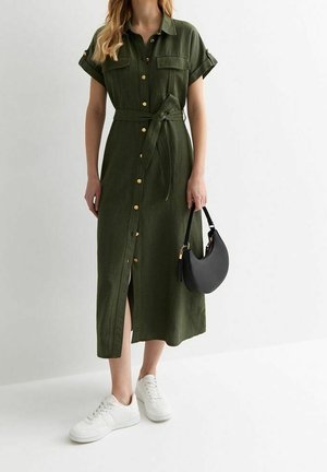 Vrouw draagt een olijfgroene midi-jurk met knopen en ceintuur in de taille, witte sneakers en houdt een zwarte halve maan handtas vast.