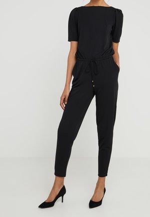 Vrouw draagt een zwarte korte mouwen jumpsuit met een koord in de taille en zwarte hoge hakken, staand tegen een lichte achtergrond.